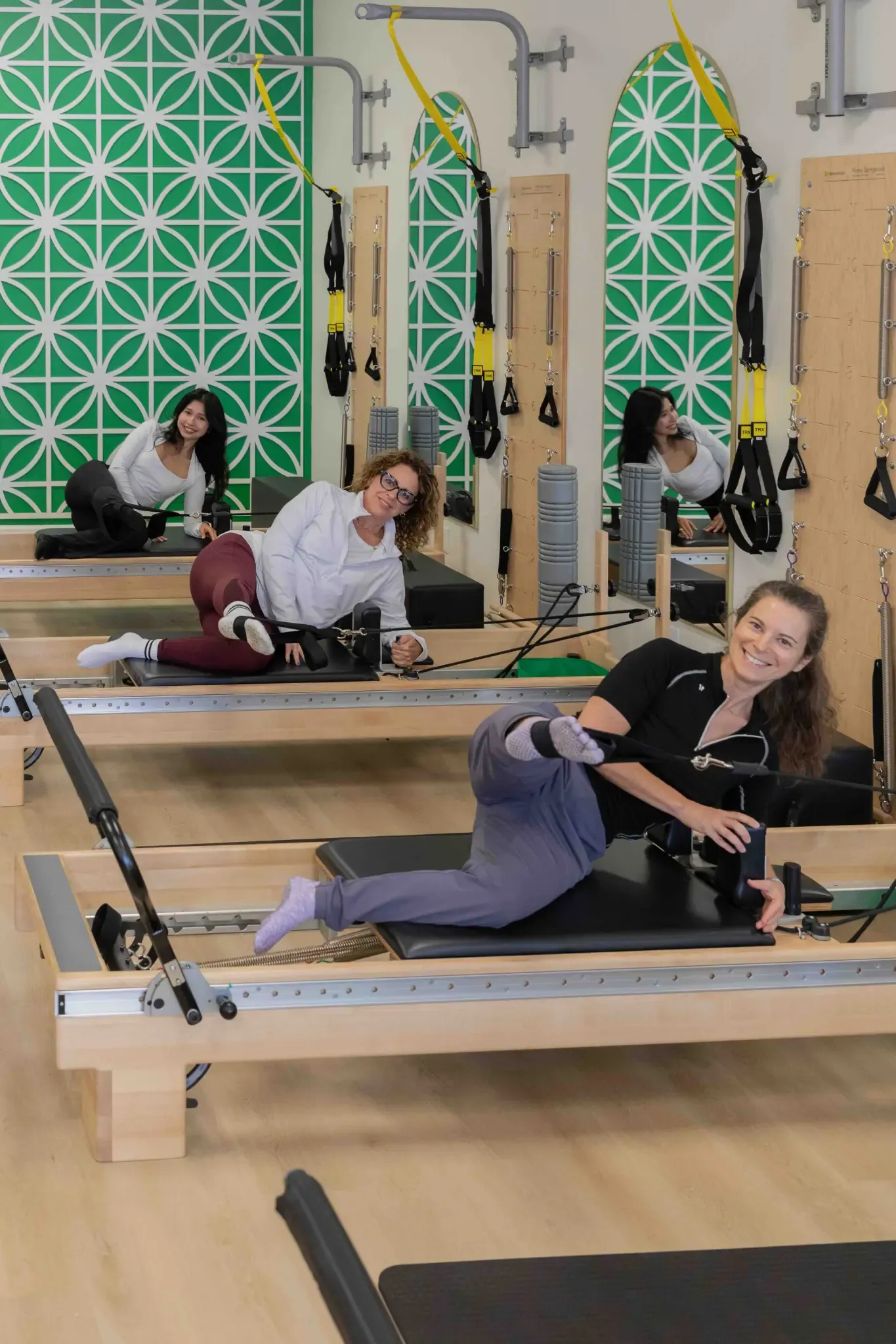 Three smiling women performing side leg exercises on reformers during a group Pilates class.