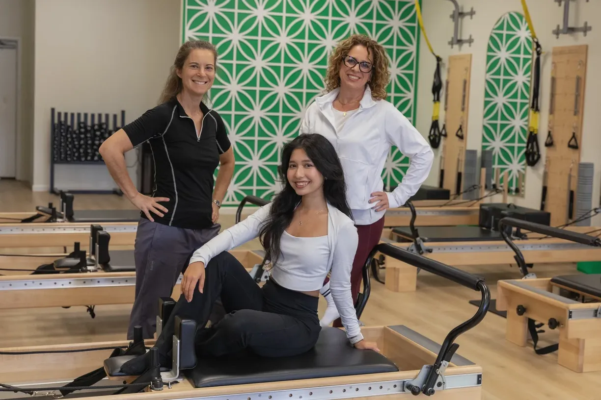 Three smiling women posing together on and around a reformer in the CharisPilates studio.